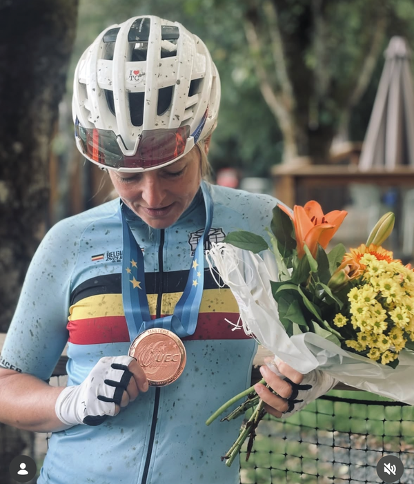 A cyclist wearing a white helmet and a light blue cycling jersey with German, Italian, and French flag details holds a bronze medal and a bouquet of orange and yellow flowers in an outdoor setting.