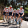 A group of cyclists riding in a peloton on a winding road surrounded by trees, wearing matching white jerseys with colorful logos and dark shorts. A black SUV is visible on the right side of the road.