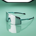A pair of shield-style sunglasses with a large mirrored lens and turquoise and black arms, photographed on a smooth mint-green surface with a reflection below. The brand name 4KAVUD is visible on the lens and arm.
