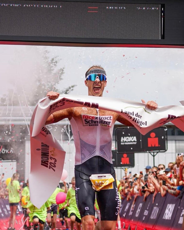 A male athlete crosses a race finish line holding a white finish banner with 'Munich 70.3' printed on it, wearing a pink and white triathlon swimsuit and black shorts, with confetti and a crowd in the background.