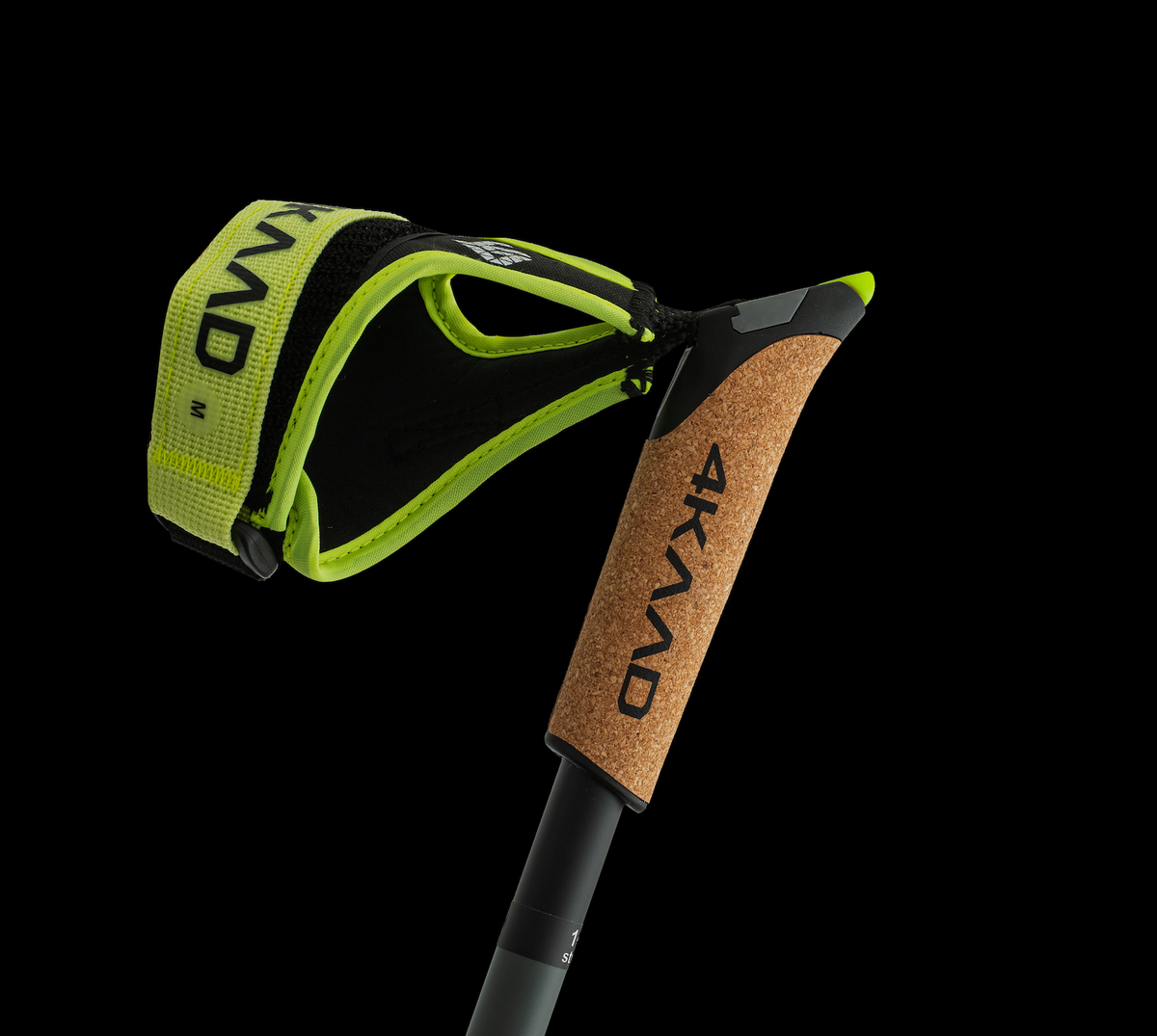 A close-up studio shot of a Nordic walking pole with a black and neon yellow-green wrist strap and a cork-textured handle grip, both branded 4KVAAD, set against a solid black background.
