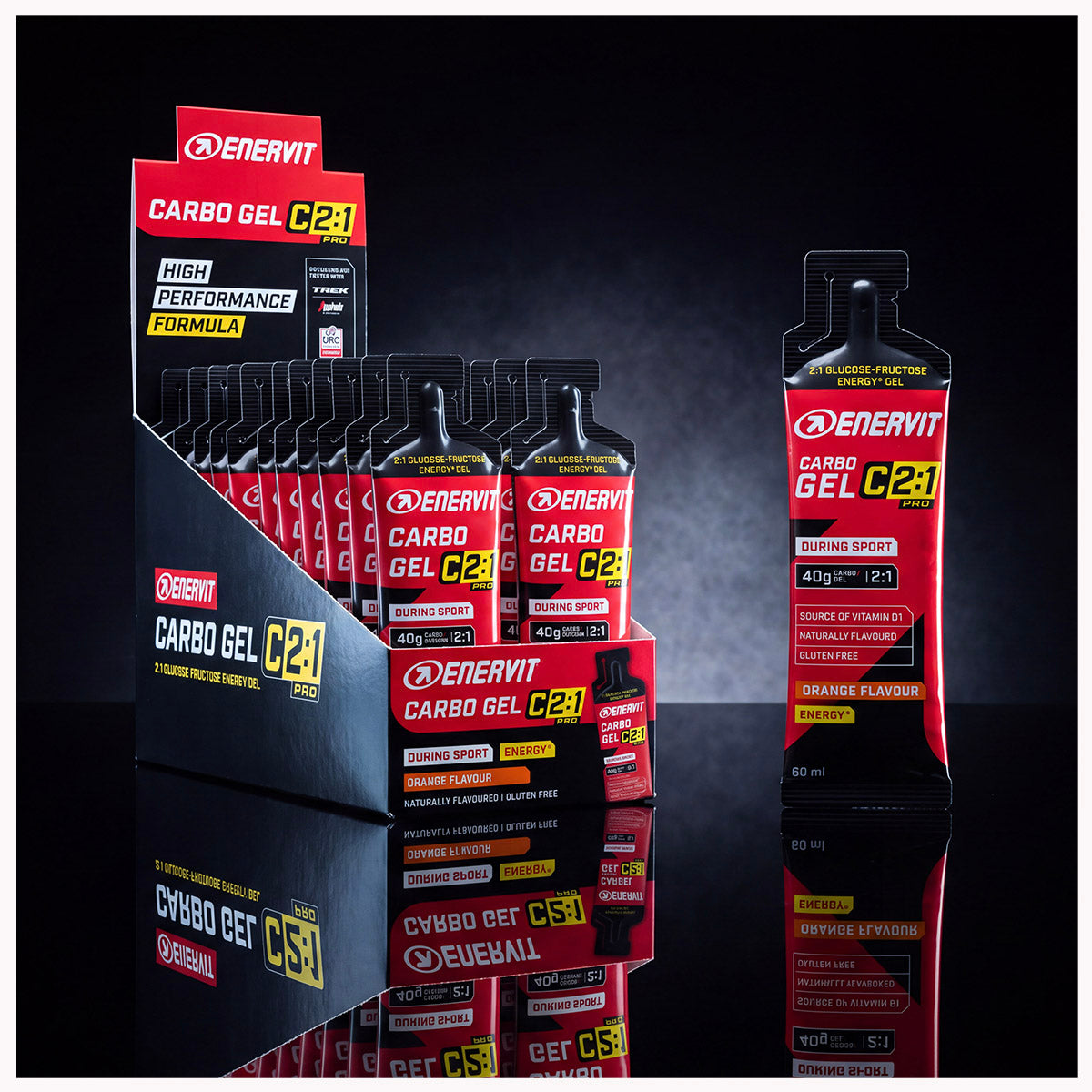 Enervit Carbogel C21 orange-flavour carbohydrate gel shown in a black retail display box with individual sachets, alongside a single larger sachet, on a dark reflective studio background.