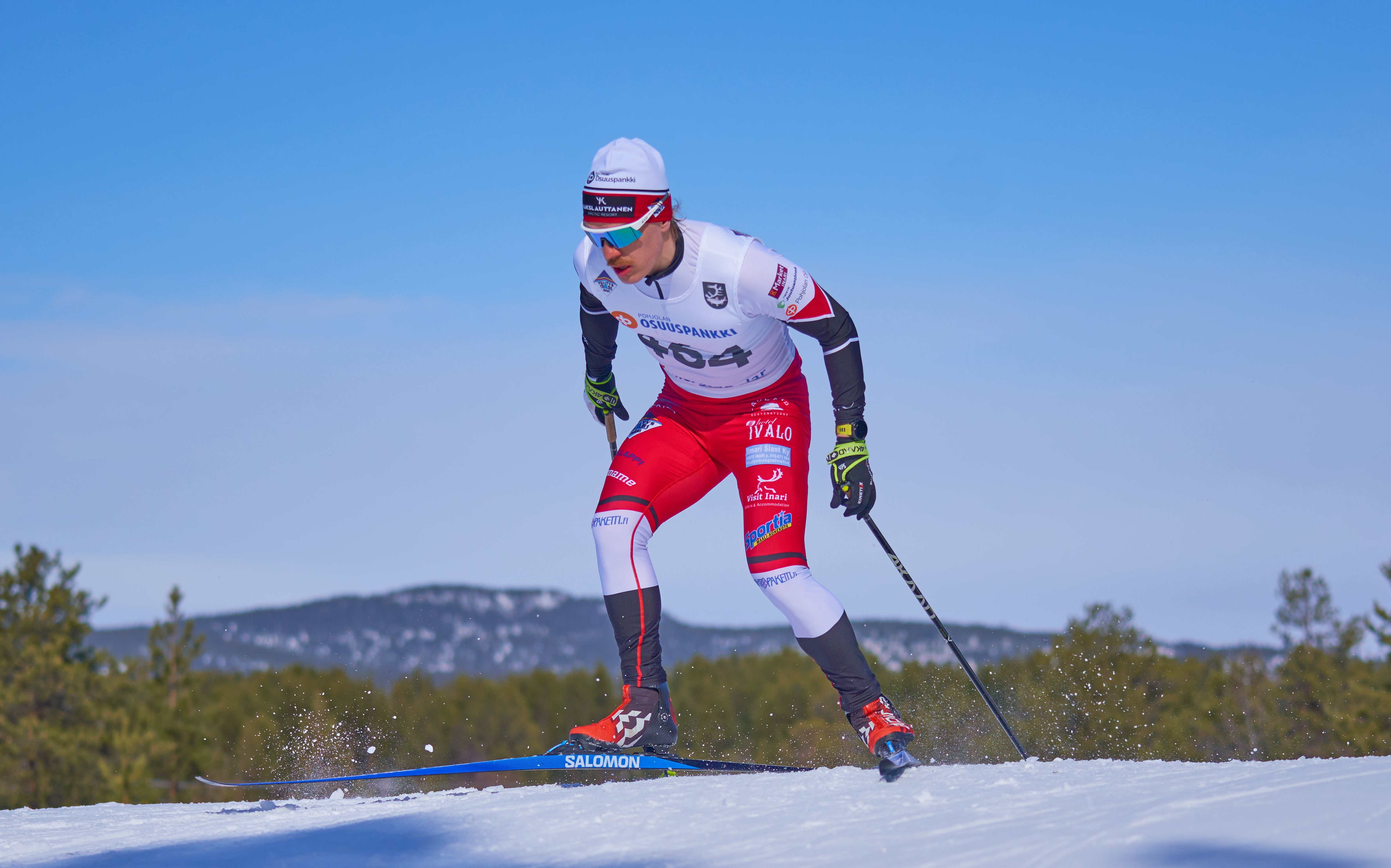 A cross-country skier in a white, red, and black racing suit glides through the snow on blue Salomon skis under a clear blue sky with distant pine trees using 4kaad carbon ski poles