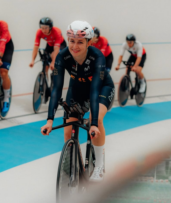 A female cyclist rides a track bicycle in a velodrome, wearing a dark navy team jersey with sponsor logos, a white helmet, and white cycling shoes. Other cyclists are visible in the background on a blue and white indoor track.