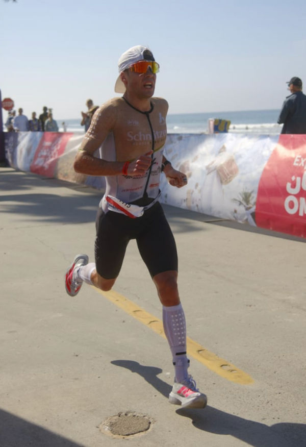 A male athlete running during a beach marathon, wearing a white and black triathlon suit, black shorts, a white cap, orange-tinted sunglasses, and white running shoes. The background shows a sunny beach course with a banner and ocean.