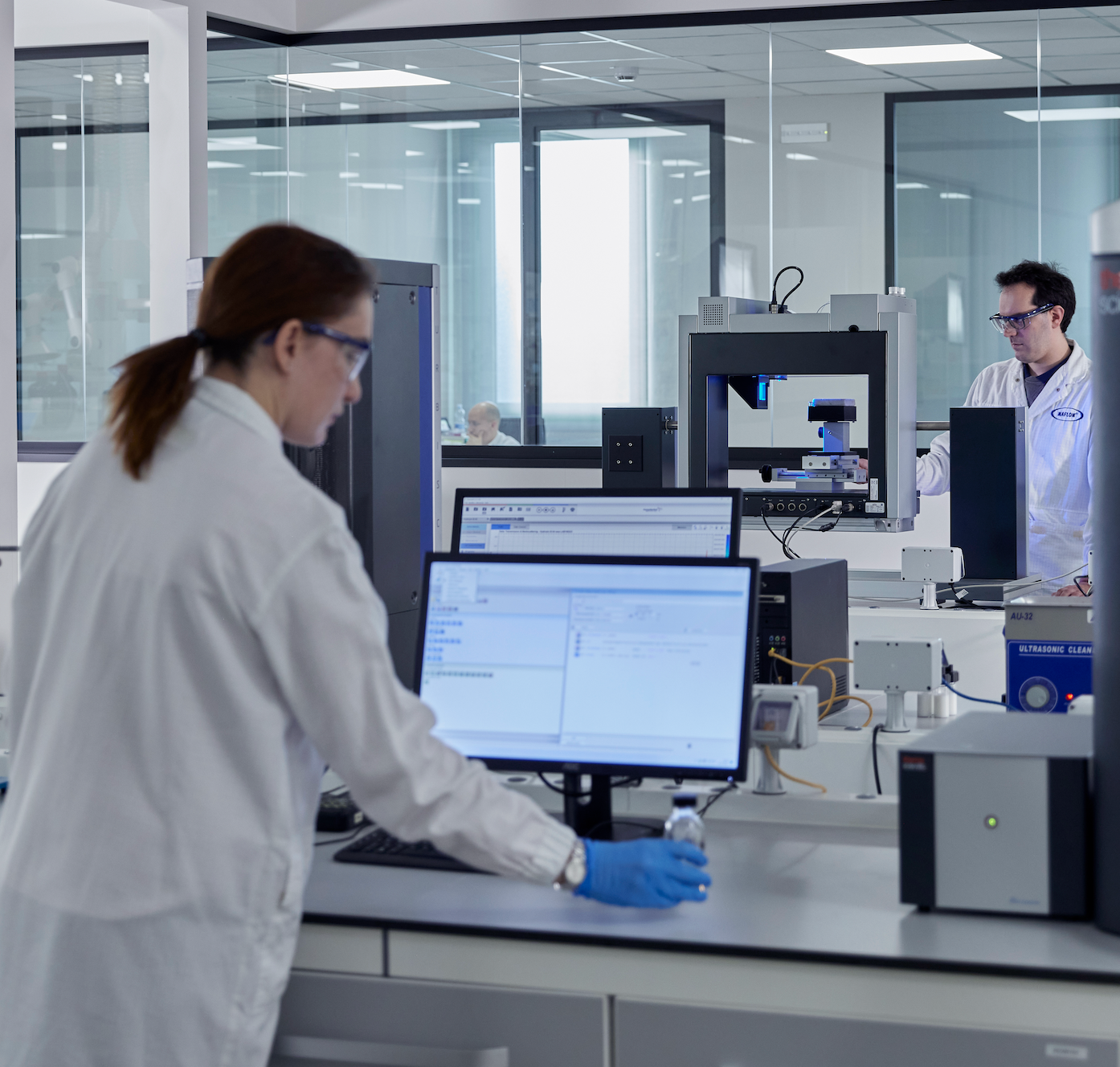 A scientist in a white lab coat and blue gloves uses a computer at a lab bench, with another scientist and laboratory equipment visible in the background. development of fluor free wax maplus brand in Finland