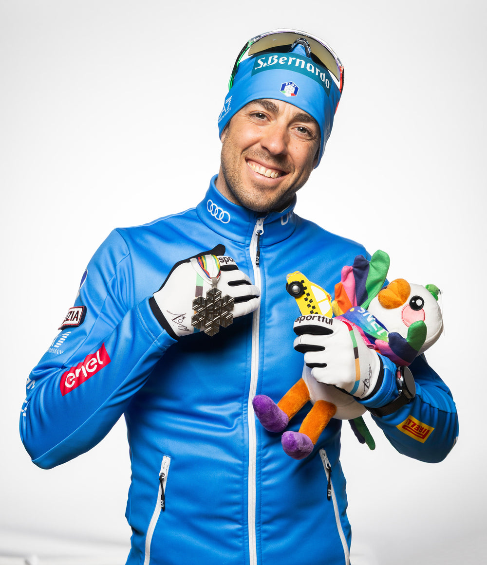 A smiling man in a bright blue ski jacket, matching blue beanie with sunglasses, and white gloves holds a colorful plush toy while displaying a ski medal. The jacket features sponsor logos including Audi, BMW, and Castore.