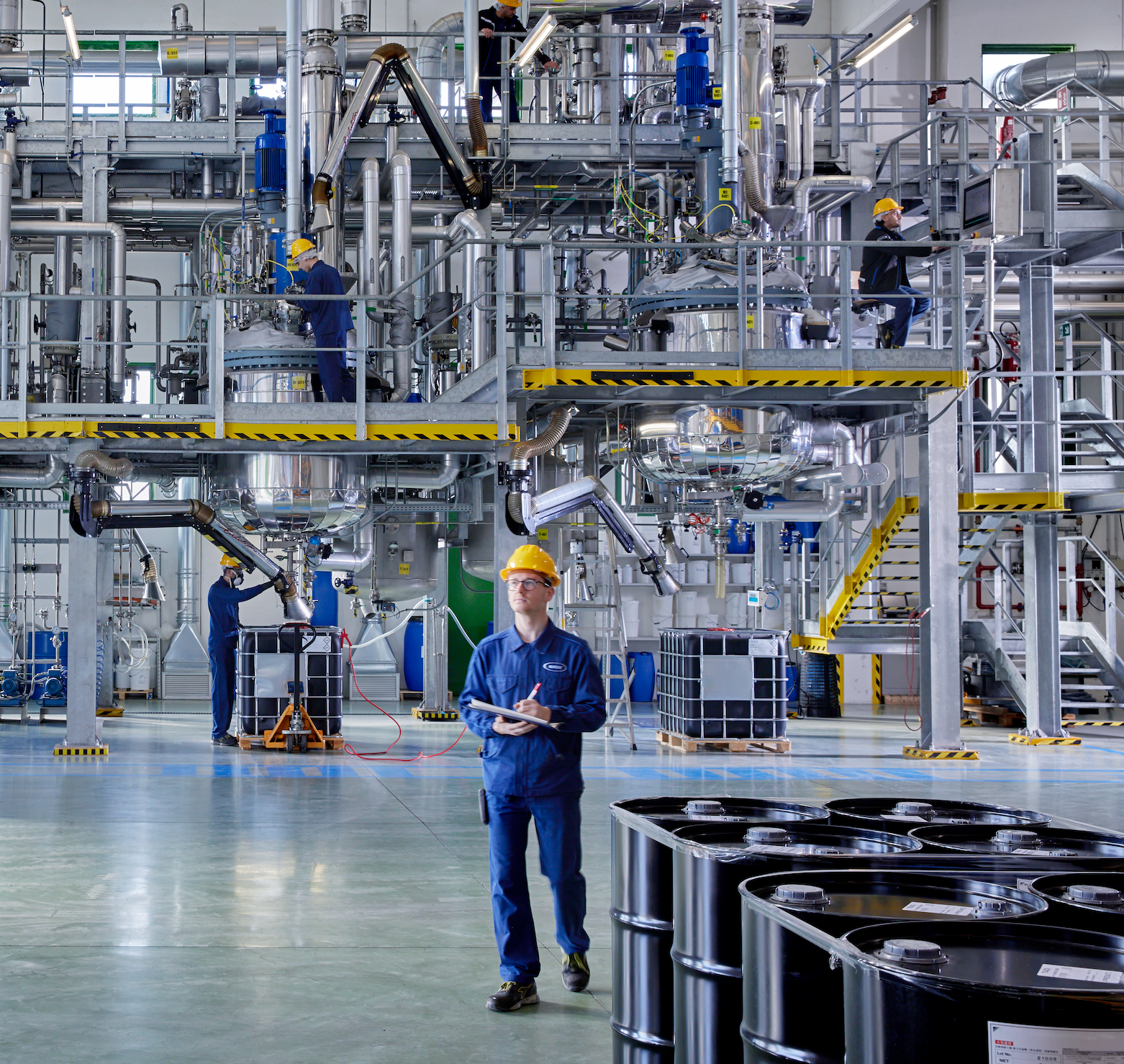 A man in a blue work uniform and yellow hard hat stands in a large industrial factory floor with metal piping, tanks, and other workers in the background. Maplus production of ski wax
