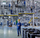 A man in a blue work uniform and yellow hard hat stands in a large industrial factory floor with metal piping, tanks, and other workers in the background. Maplus production of ski wax