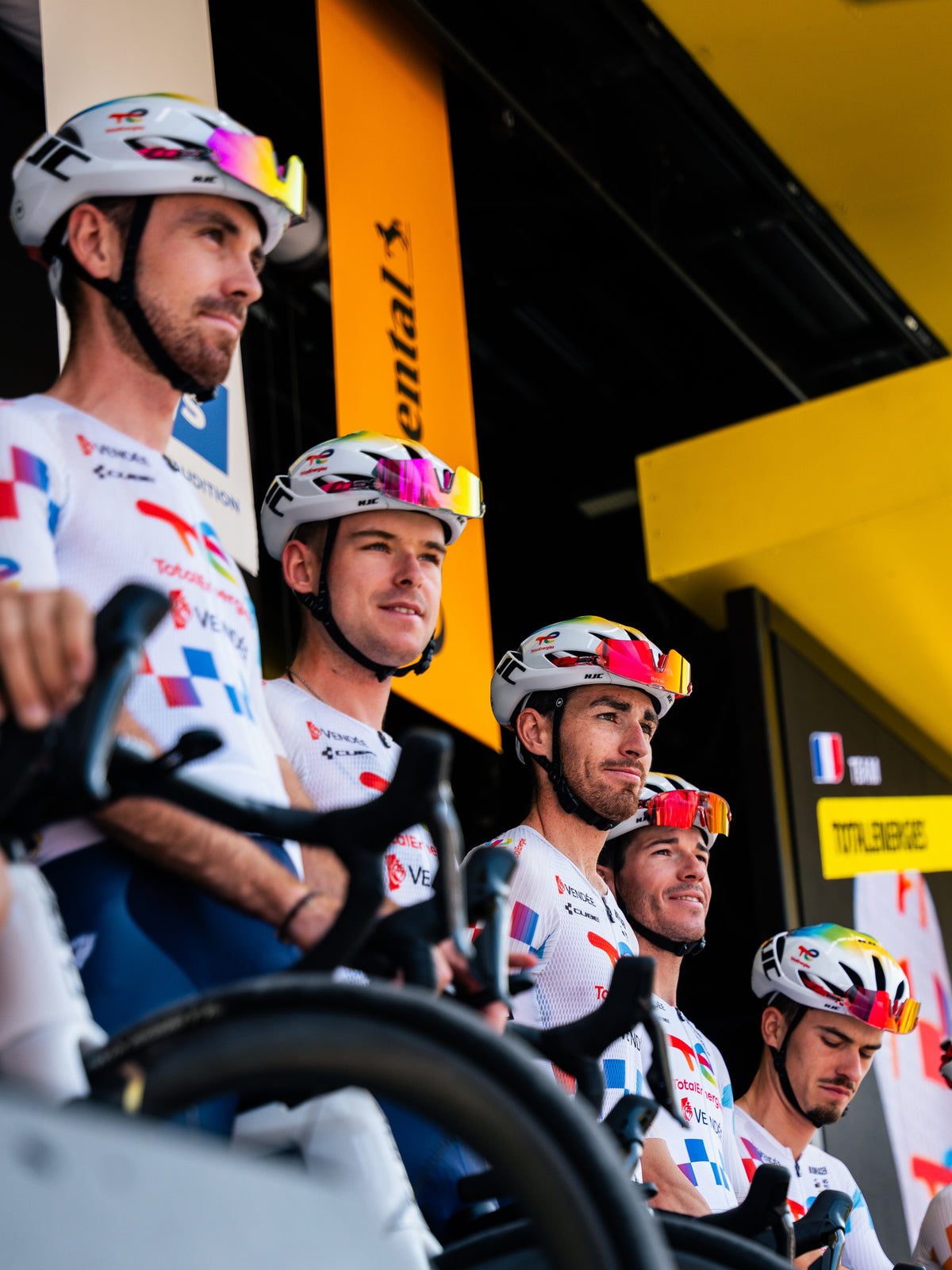 A group of professional cyclists wearing white team jerseys with red and blue graphics, white helmets, and colorful sunglasses, riding bicycles in a bright cycling team environment. 4kaad sports glasses used by every rider at tour de france