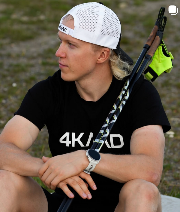 A man wearing a black 4KAAD t-shirt and a white mesh-back cap sits outdoors with a pair of trail running shoes resting on his shoulder. He is wearing a smartwatch and a bright neon green wrist strap.