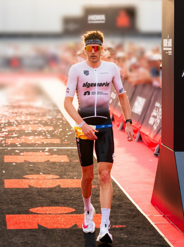 A male athlete walking on a red carpet after a race, wearing a white and black gradient cycling jersey with 'algenapin' printed on the chest, black shorts, and white running shoes.