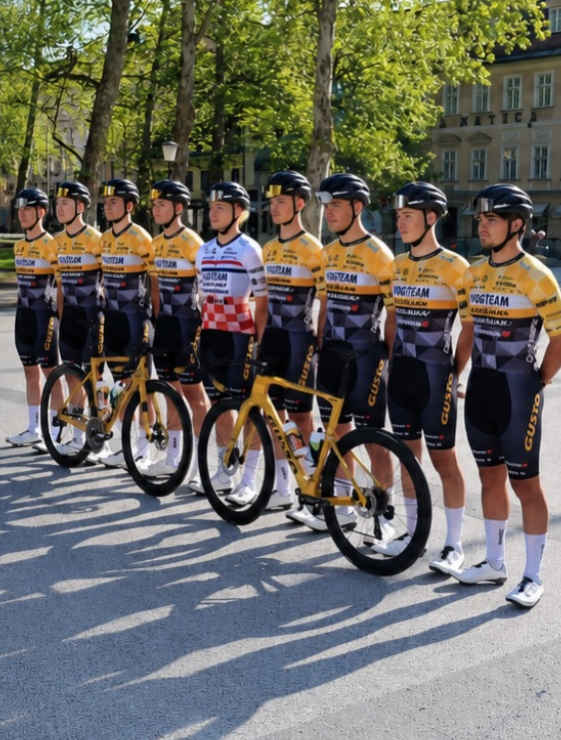 A group of cyclists standing outdoors on a sunny day with trees and a building in the background. Most wear matching yellow, black, and gray cycling jerseys and shorts, with one rider in a white and red checkered jersey.