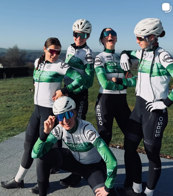 Five cyclists posing outdoors in matching white, green, and black SERSO cycling jerseys, helmets, sunglasses, and bib shorts on a paved path with grass and trees in the background.