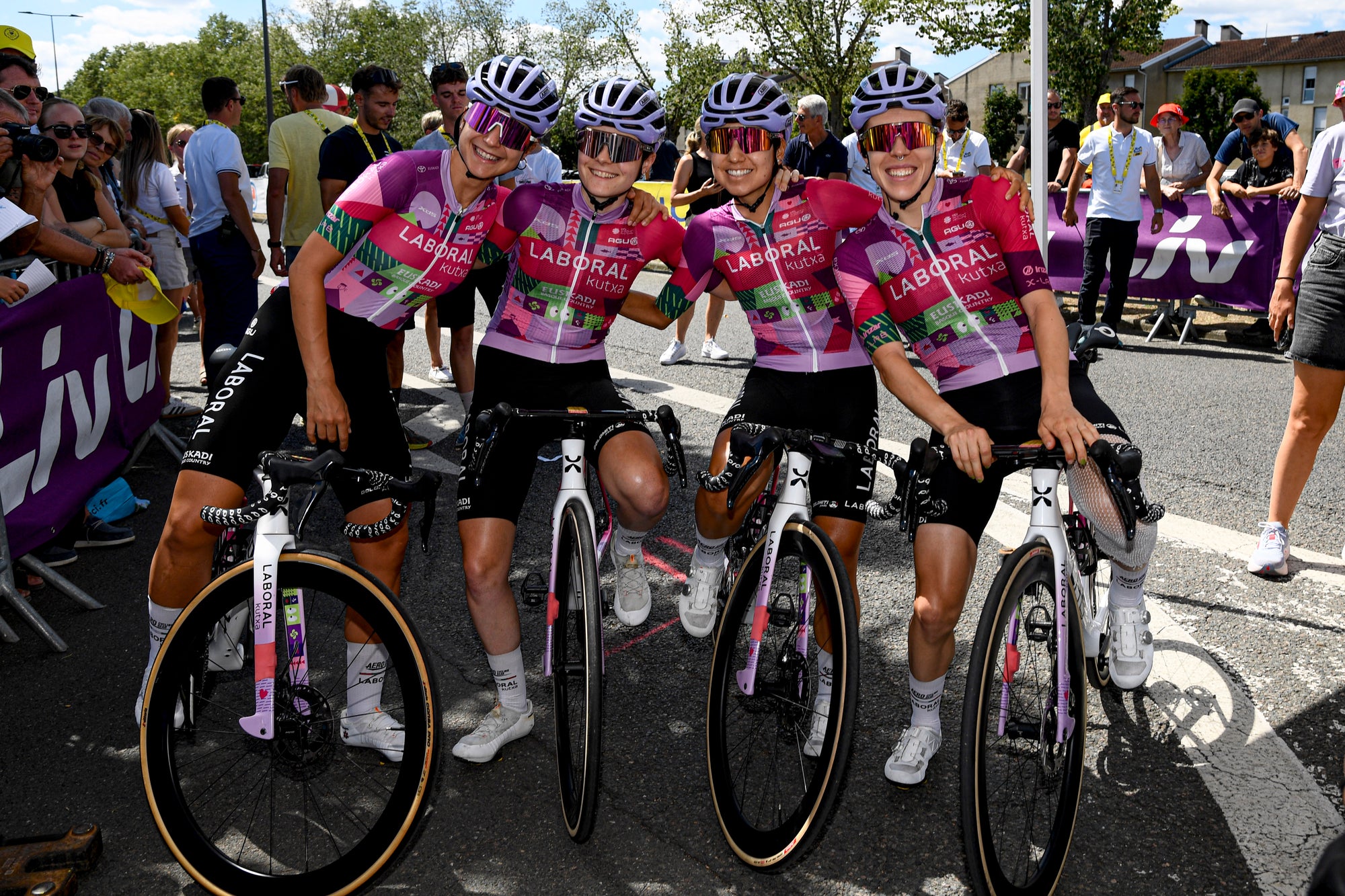 Four female cyclists in matching pink and purple Liberaal team jerseys pose with their road bikes on a street during a race, with spectators and barriers in the background. 4kaad eyewear by team laboral kutxa spain