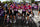 Four female cyclists in matching pink and purple Liberaal team jerseys pose with their road bikes on a street during a race, with spectators and barriers in the background. 4kaad eyewear by team laboral kutxa spain