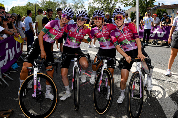 Four female cyclists in matching pink and purple Liberaal team jerseys pose with their road bikes on a street during a race, with spectators and barriers in the background. 4kaad eyewear by team laboral kutxa spain