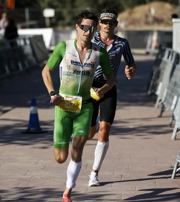 Two male runners during a road race. The runner in the foreground wears a green and white Castore cycling jersey and shorts, white knee sleeves, and sunglasses.