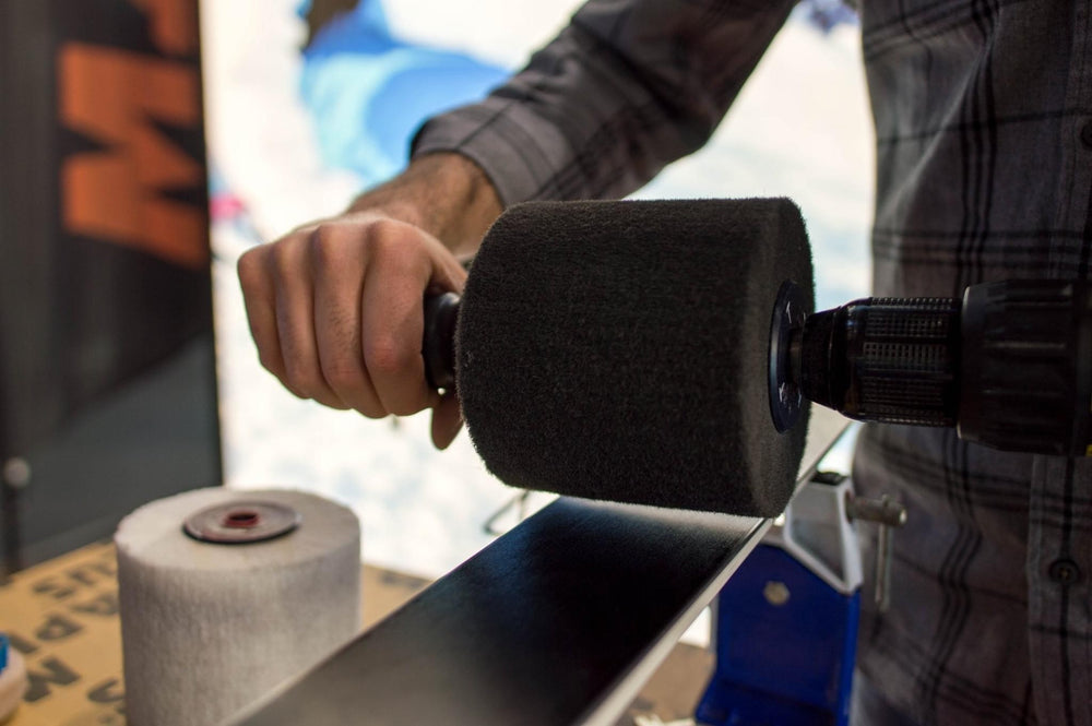 A close-up action shot of a person using a foam-covered drill attachment to work on a long metal rod or rail, with a spool of material nearby and a blurred outdoor background.