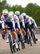 A group of professional cyclists riding in a peloton on an asphalt road, wearing matching white and navy cycling kits with various sponsor logos.