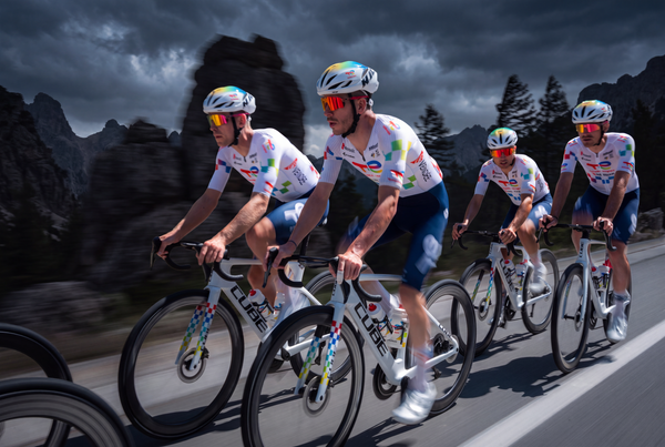 Four cyclists riding road bikes in a peloton on a mountain road under a dark, dramatic sky. They wear matching white jerseys with multicolor accents, blue shorts, and white helmets. 4Kaad eyewear supplier Team Total energies