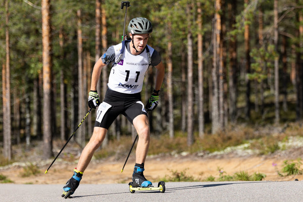 A male athlete in winter sports gear, including a helmet, bib number 11, and ski boots, actively skiing on a paved road surrounded by a dense pine forest.