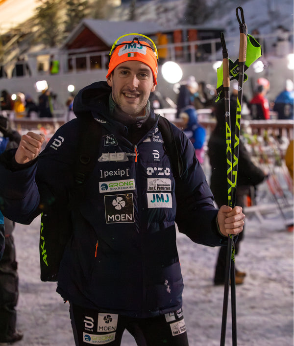 A male skier wearing a dark navy sponsor jacket, black bib pants, and an orange beanie with a green and white flag, holding ski poles in a snowy outdoor setting at dusk.