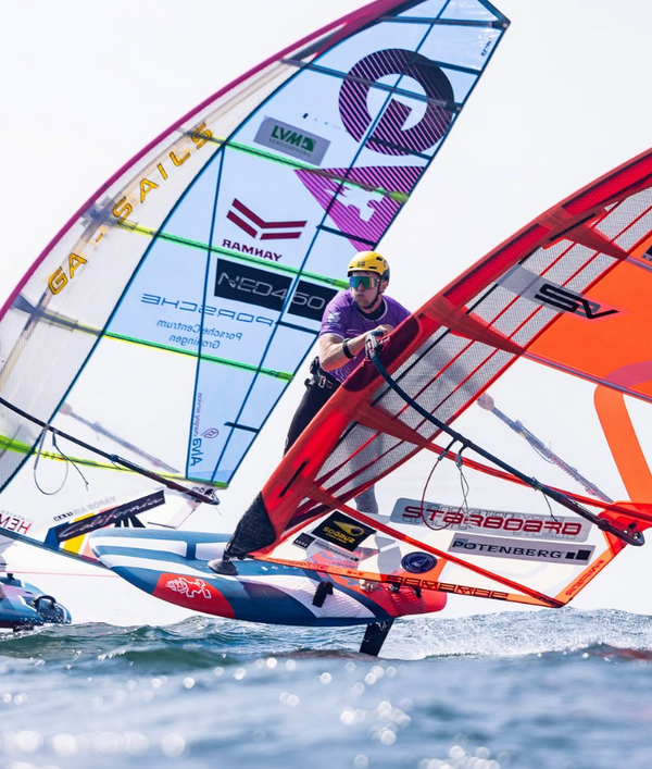 A windsurfer in a purple top and helmet maneuvers a red, white, and blue foil surfboard with a bright orange sail over open water. Another competitor's sail is visible in the background under a bright sky.